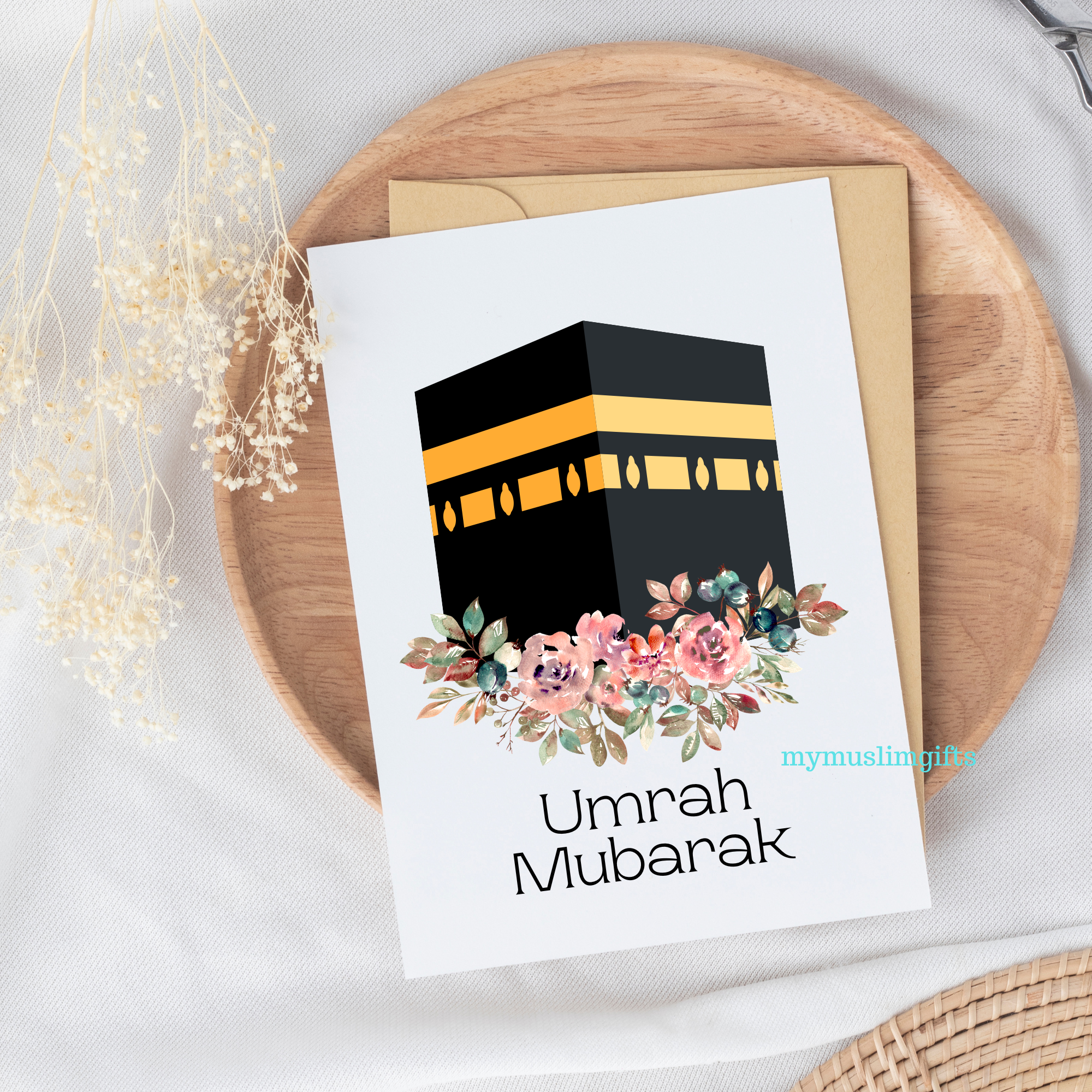 A greeting card with a image of the Kaaba and floral design, accompanied by the text 'Umrah Mubarak'. The card is placed on a wooden surface with a neutral background.